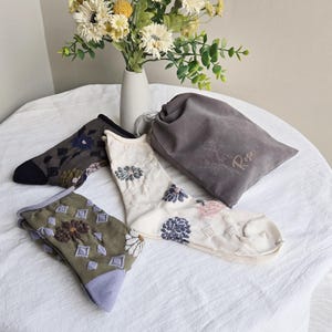 May include: Three pairs of patterned socks with floral embroidery and a gray drawstring bag. One pair is olive green with purple accents, another is white with blue and pink flowers, and the third is dark gray with blue flowers.