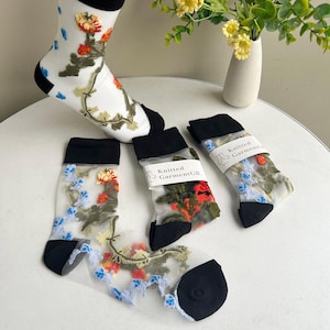 Pode incluir: Três pares de meias transparentes bordadas com flores com elásticos pretos. As meias são brancas com bordados florais coloridos. As meias têm uma etiqueta que diz "Knitted GarmentGB."