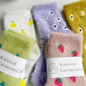 Könnte beinhalten: Nahaufnahme von mehreren Paaren flauschiger Socken in verschiedenen Farben, darunter Weiß, Grün, Lavendel, Rosa und Gelb. Die Socken zeigen Fruchtmuster wie Zitronen, Erdbeeren und Birnen. Jedes Paar hat ein weißes Etikett mit der Aufschrift "Knitted Garment GB."