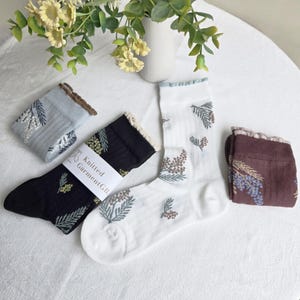 Op de afbeelding: Een assortiment sokken in verschillende kleuren, waaronder zwart, wit, blauw en bruin. De sokken hebben een bloemenpatroon en ruches. Eén paar is verpakt met een label met de tekst "Knitted Garment GB".