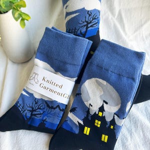 Op de afbeelding: Drie paar sokken met Halloween-thema. De sokken hebben een blauw en zwart kleurenschema met afbeeldingen van een spookhuis, vleermuizen en een volle maan. De tekst "Knitted GarmentGB" is zichtbaar.