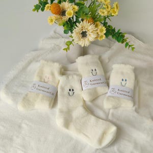 May include: Four pairs of cream-coloured fluffy socks with embroidered smiley faces. Each pair has a white band with the text "Knitted GarmentGB". A vase of yellow flowers is in the background.