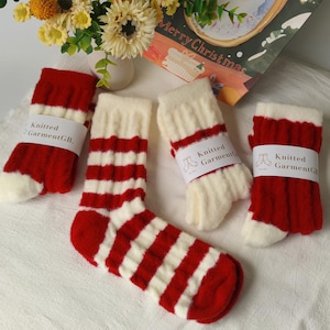 May include: Four pairs of red and white striped knit socks. One pair is fully displayed, while the others are rolled up and wrapped with a white label that reads "Knitted GarmentGB." The socks have white toes and cuffs. A "Merry Christmas" card is in the background.