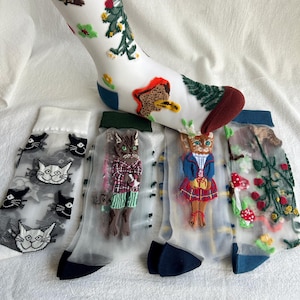 May include: Four pairs of sheer socks with embroidered cat designs. The socks are white with black trim and feature different cat illustrations, including a cat wearing a plaid shirt, a cat wearing a blue dress, and a cat with a red bow.