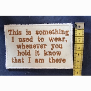 Könnte beinhalten: Ein weißer Stoff-Patch mit brauner Stickerei, auf dem steht "This is something I used to wear, whenever you hold it know that I am there". Der Patch befindet sich neben einem gelben Maßband.