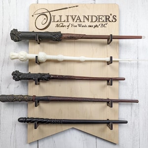 May include: A wooden wand display rack with the text "Ollivanders* Makers of Fine Wands since 382 BC."  Five wands are displayed on the rack, including a brown wand, a white wand, a black wand, a brown wand with gold accents, and a black wand with silver accents.