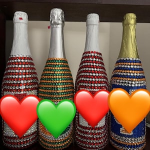 May include: Four champagne bottles decorated with colorful rhinestones. The bottles feature red, green, and blue rhinestone patterns. Each bottle has a silver foil top.