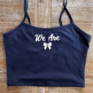 May include: Navy blue crop top with thin straps. The top features the white text "We Are" above a white bow graphic. The top is made of a soft material.