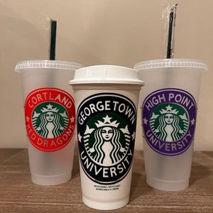 May include: Three clear plastic cups with lids and straws. Each cup has a different university logo printed on it. The logos are for Cortland Red Dragons, Georgetown University, and High Point University.