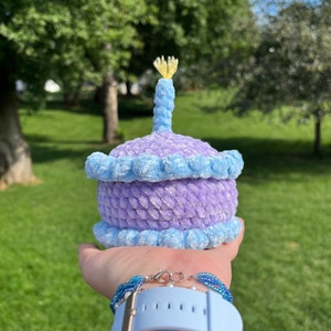 May include: A purple and blue crocheted cake with a yellow candle on top. The cake is being held in someone's hand.
