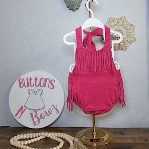 May include: A fuchsia-colored romper with fringe detailing, displayed on a white hanger. The romper has a halter neck and a bow at the back. A wooden sign with the text "Buttons N Bowz" is in the image.