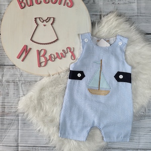 May include: A blue and white striped baby onesie with a sailboat applique. The onesie has a navy blue trim and white buttons. The onesie is on a white fur background.