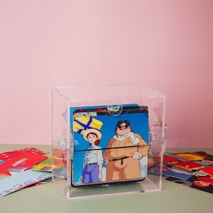 Può includere: Una scatola in acrilico trasparente con un display rotante di carte da collezione di personaggi dei cartoni animati colorati. Le carte mostrano una ragazza con un cappello giallo e un uomo con un naso da maiale e occhiali da aviatore. La scatola è su una superficie verde con un mucchio di altre carte da collezione.