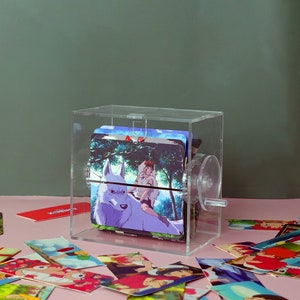 Puede incluir: Una caja de acr&iacute;lico transparente con un mecanismo giratorio, que muestra una pila de tarjetas coloridas con personajes de anime. Las tarjetas representan un lobo blanco y una persona montando al lobo. La caja est&aacute; rodeada de tarjetas dispersas con im&aacute;genes similares.