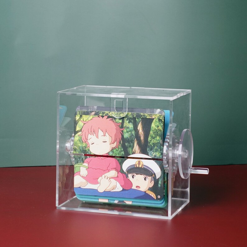 May include: A clear acrylic box with a rotating mechanism, displaying a series of colorful cards featuring cartoon characters. The cards depict a girl with pink hair and a boy wearing a sailor hat.