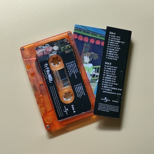 Pu&ograve; includere: Una cassetta audio arancione con un'etichetta nera che presenta il testo giapponese "千と千尋の神隠し" (La citt&agrave; incantata) e un'immagine di Chihiro dal film. La cassetta audio &egrave; accanto a un inserto di carta bianco e nero con la lista delle tracce dell'album.