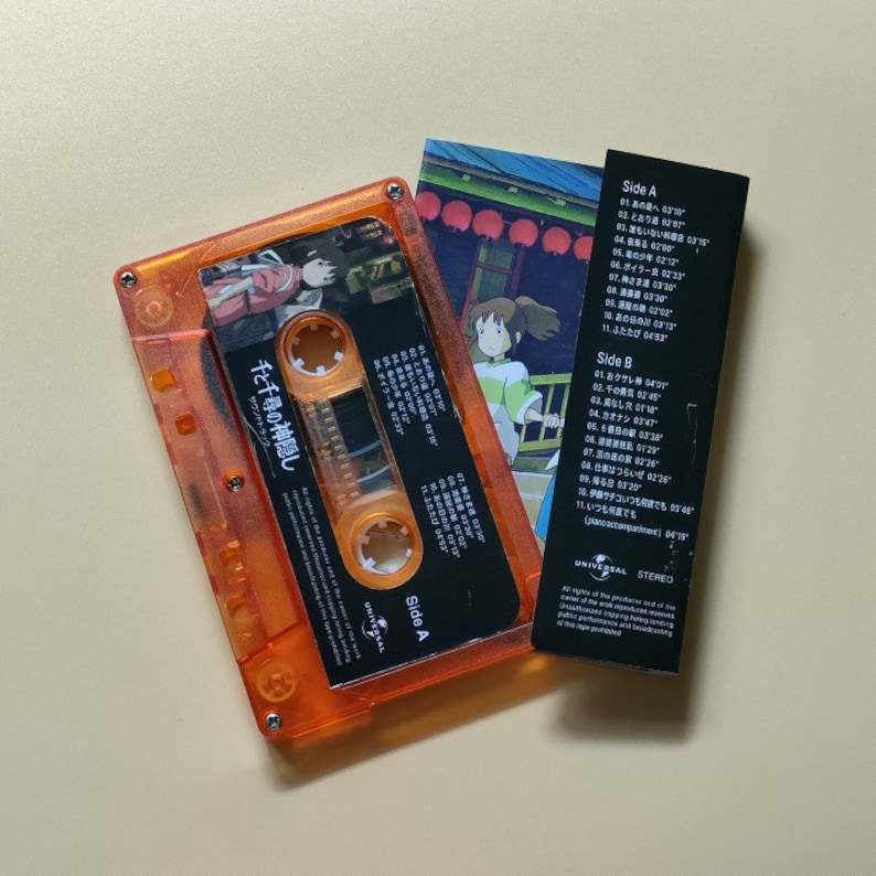 Pu&ograve; includere: Una cassetta audio arancione con un'etichetta nera che presenta il testo giapponese "千と千尋の神隠し" (La citt&agrave; incantata) e un'immagine di Chihiro dal film. La cassetta audio &egrave; accanto a un inserto di carta bianco e nero con la lista delle tracce dell'album.