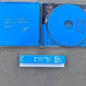 May include: A blue CD case with a blue CD inside. The CD case has a handwritten message on the back that says "久石こくやりましたね! 全部ミニマルですなんて、ありカレーとう" and a signature. The CD has the text "君たちは 生きろか どう サウンドトラック" on it.