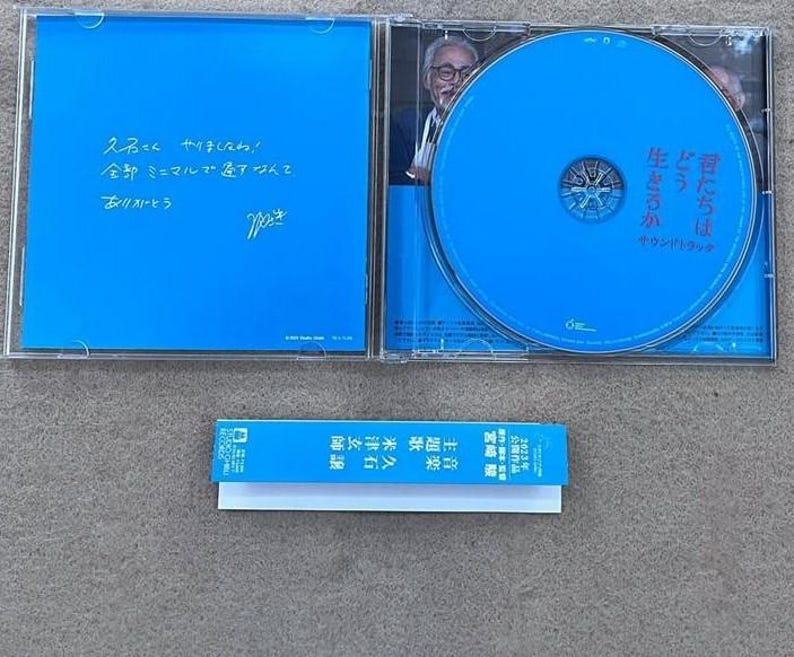 May include: A blue CD case with a blue CD inside. The CD case has a handwritten message on the back that says "久石こくやりましたね! 全部ミニマルですなんて、ありカレーとう" and a signature. The CD has the text "君たちは 生きろか どう サウンドトラック" on it.