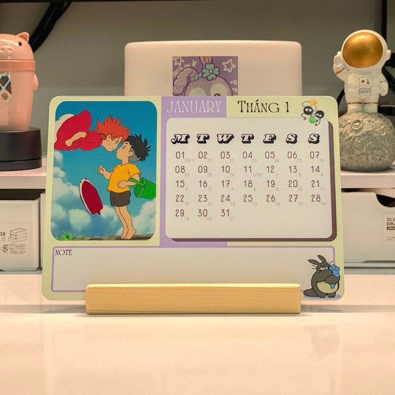 May include: A colorful desk calendar with a cartoon image of two characters from the animated film 'Kiki's Delivery Service'. The calendar is for January and has the Vietnamese word 'Th&aacute;ng 1' for 'Month 1' printed on it. The calendar has a wooden stand.
