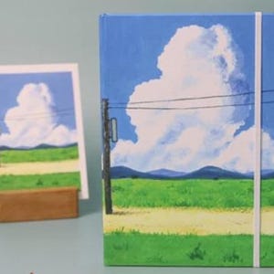 May include: A blue and green landscape notebook with a white cloud and power lines. The notebook has a white elastic band closure.