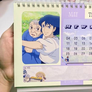 Może przedstawiać: Spiralnie oprawiony kalendarz z majową stroną. Zawiera ilustrację w stylu anime z dwoma postaciami. Kalendarz ma jasnozieloną obw&oacute;dkę i fioletową sekcję z dniami tygodnia. Na dole mała ilustracja.