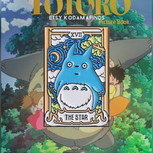 May include: A blue and white enamel pin featuring Totoro from the movie My Neighbor Totoro. The pin is in the style of a tarot card, with the number XVII and the words "The Star" on it. The pin is surrounded by a gold frame.