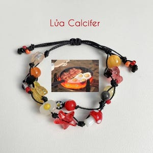 May include: A black string bracelet with various colourful beads, including red, yellow, orange, and white. The bracelet features a charm of a frying pan with eggs and bacon, a mushroom, a star, and a flower. The text "Lúa Calcifer" is written in red above the bracelet.