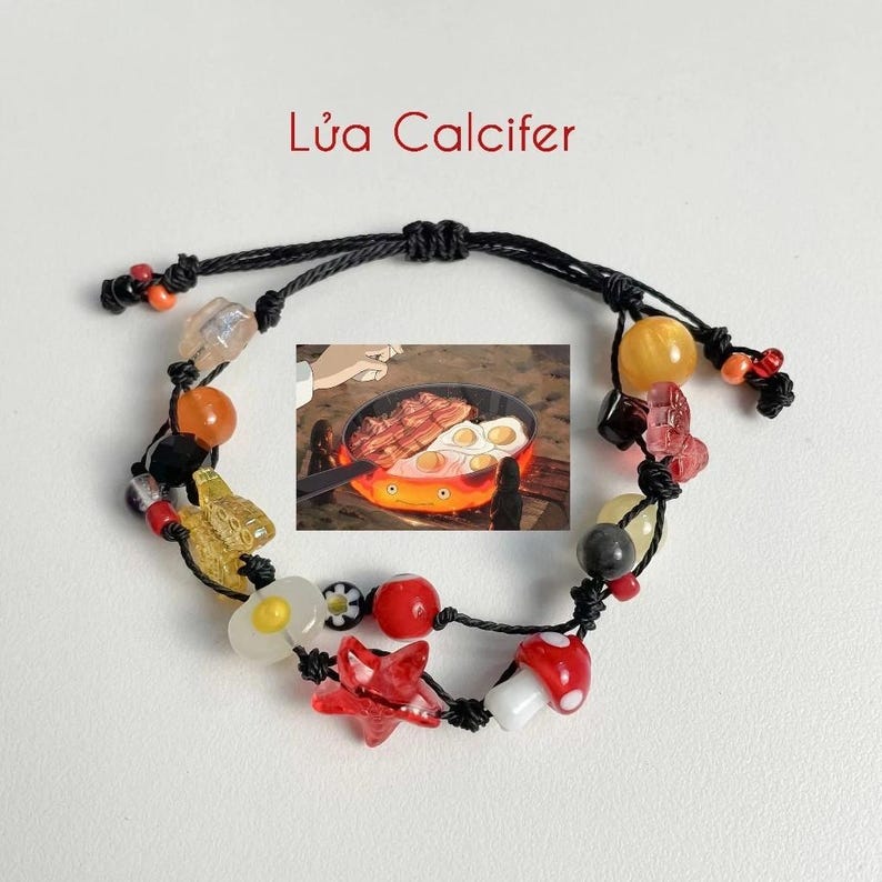 Puede incluir: Un brazalete de cuerda negra con varias cuentas de colores, incluyendo rojo, amarillo, naranja y blanco. El brazalete presenta un dije de una sart&eacute;n con huevos y tocino, un hongo, una estrella y una flor. El texto "L&uacute;a Calcifer" est&aacute; escrito en rojo sobre el brazalete.