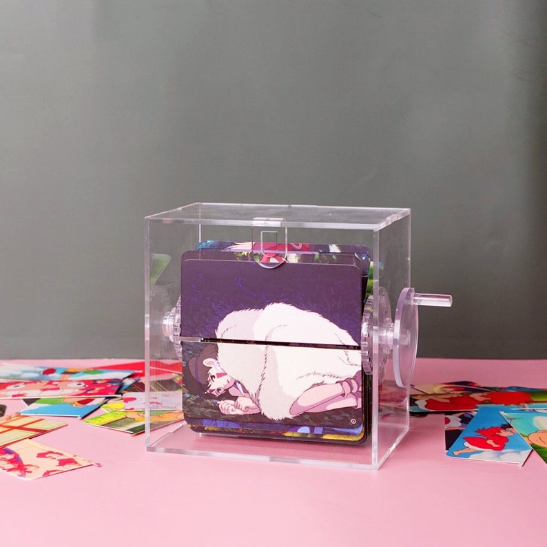 Puede incluir: Un porta tarjetas de acr&iacute;lico transparente con una manivela en el lateral. El porta tarjetas est&aacute; lleno de tarjetas con personajes de anime. Las tarjetas son visibles a trav&eacute;s del acr&iacute;lico transparente.