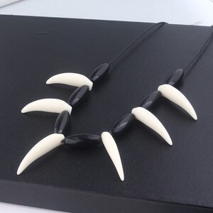 May include: Black and white necklace with a series of black beads and white plastic fangs. The necklace is on a black surface.