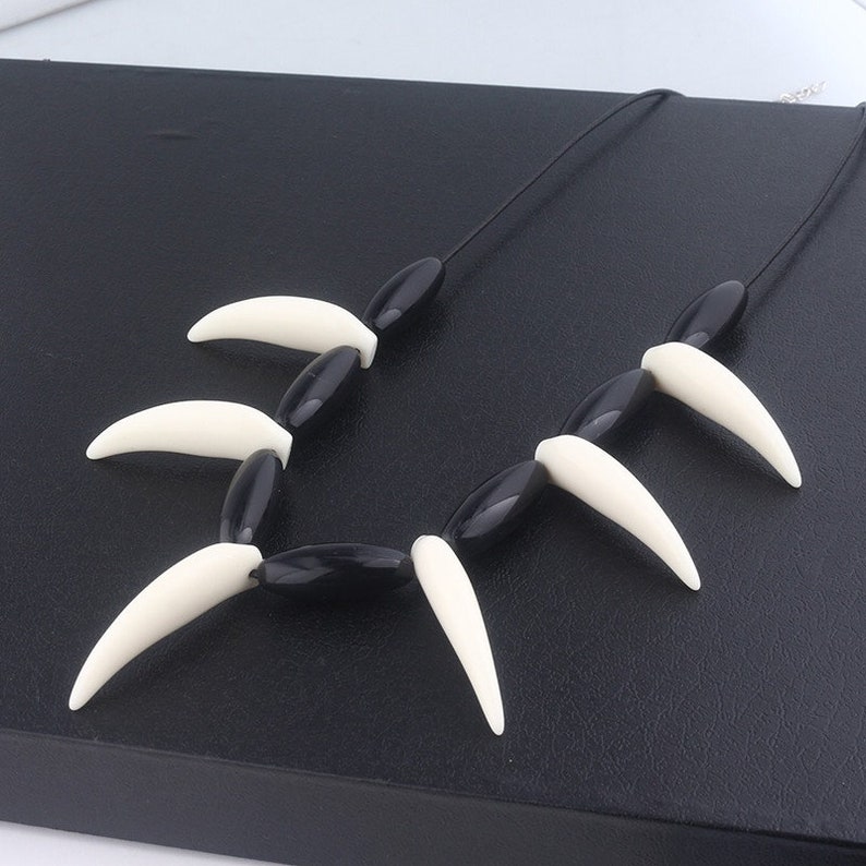 May include: Black and white necklace with a series of black beads and white plastic fangs. The necklace is on a black surface.