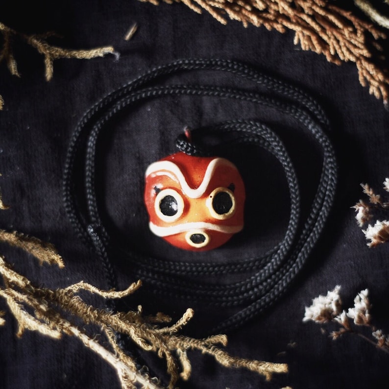 May include: A red and white No Face mask pendant necklace with black eyes and a black mouth. The necklace is on a black cord and is lying on a dark fabric background.