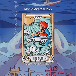 May include: A gold and blue enamel pin featuring a character from the animated film "Ponyo" sitting on a boat with a shark fin in the water. The pin is a depiction of the "The Sun" card from the tarot deck. The text "生まれてきてよかった" is written in Japanese on the left side of the pin. The text "The Sun" and "呂崎駿監督作品" are written in English and Japanese on the bottom of the pin.