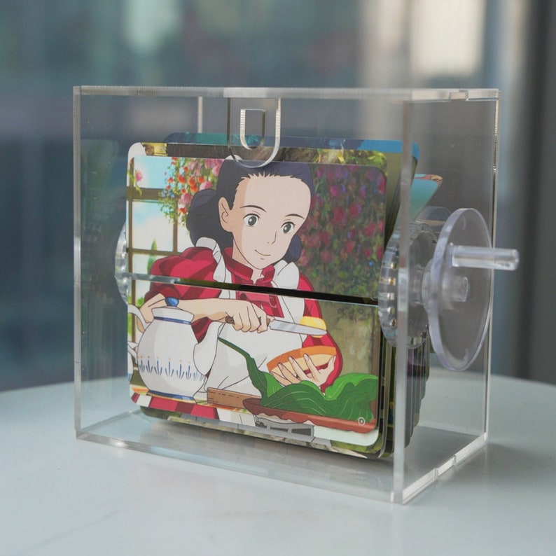 K&ouml;nnte beinhalten: Eine transparente Acrylbox mit einem Drehmechanismus, die eine Reihe von bunten Karten zeigt, auf denen ein Anime-Charakter Essen zubereitet. Die Karten zeigen eine Szene aus einer K&uuml;che mit einer Teekanne, einem Messer und einem Teller mit Essen.