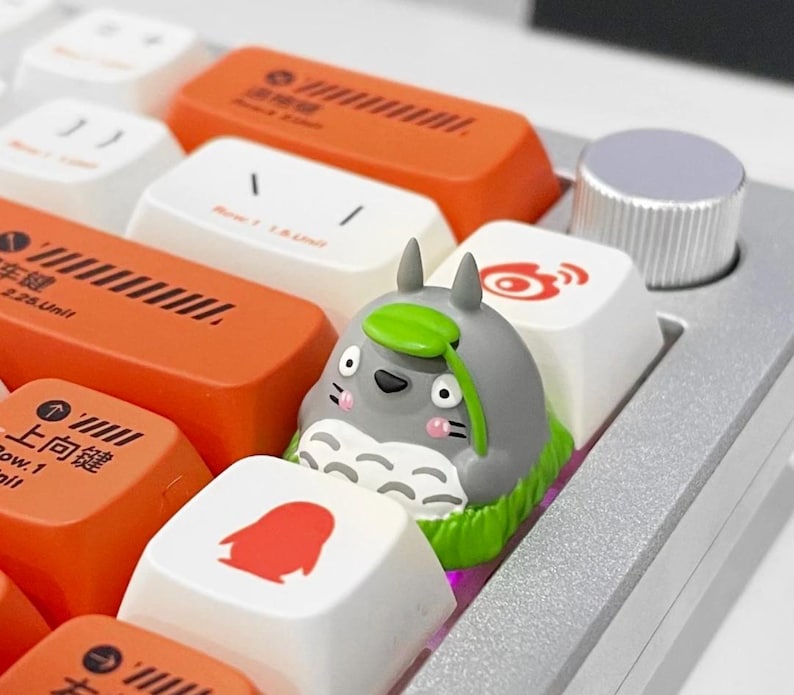 May include: A close-up of a mechanical keyboard with custom keycaps. The keycaps are a mix of white and orange, with a gray Totoro keycap in the center. The keyboard also has a silver volume knob.
