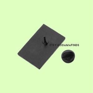 May include: Black enamel pin with a rectangular shape and a silver pin back. The pin is on a light green background.