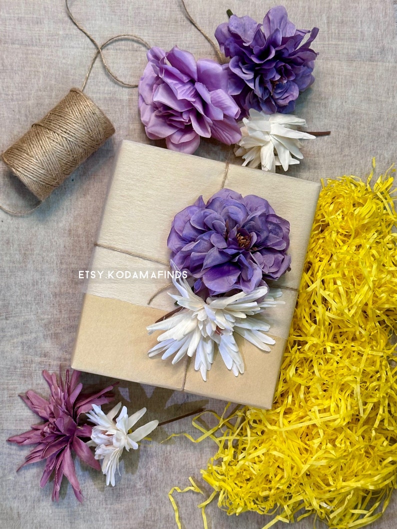 May include: A brown gift box wrapped with twine and decorated with purple and white flowers. The box is surrounded by yellow shredded paper.