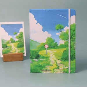 May include: A blue and green landscape painting on a notebook cover. The painting depicts a winding dirt path leading up a hill with a red stop sign. The sky is blue with white clouds and a small plane flying overhead.
