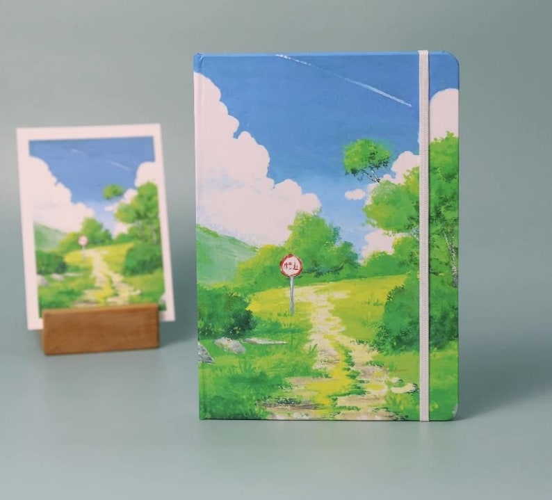 May include: A blue and green landscape painting on a notebook cover. The painting depicts a winding dirt path leading up a hill with a red stop sign. The sky is blue with white clouds and a small plane flying overhead.