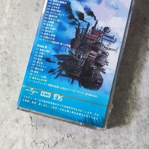 Pu&ograve; includere: Una custodia per cassette con sfondo blu e un'immagine di una casa volante. La custodia &egrave; per la colonna sonora del film "Il castello errante di Howl". Il testo sulla custodia recita "Il castello errante di Howl - Un giro di giostra della vita".