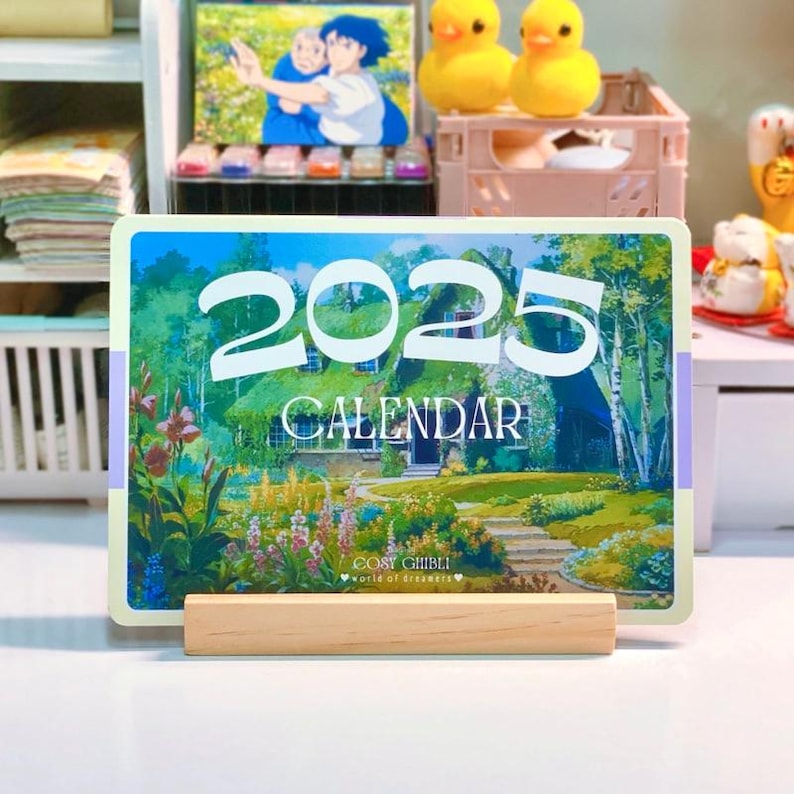 May include: A 2023 calendar featuring a whimsical illustration of a cottage with a thatched roof, surrounded by lush greenery and flowers. The text "COSY GHIBLI" and "World of Dreams" is printed at the bottom of the calendar.
