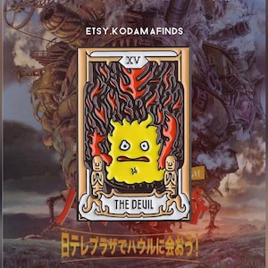 May include: A gold-colored enamel pin featuring a yellow cartoon character with a red and black flame design. The pin is a depiction of the Devil card from a tarot deck. The text "XV THE DEUIL" is printed on the pin.
