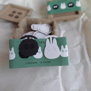 May include: A green card with the text "2 designs 16 pieces" and the word "clips" printed on it. The card features two white felt cutouts of a cartoon character with large eyes and a black felt cutout of a cartoon character with large eyes.