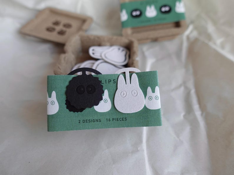 May include: A green card with the text "2 designs 16 pieces" and the word "clips" printed on it. The card features two white felt cutouts of a cartoon character with large eyes and a black felt cutout of a cartoon character with large eyes.