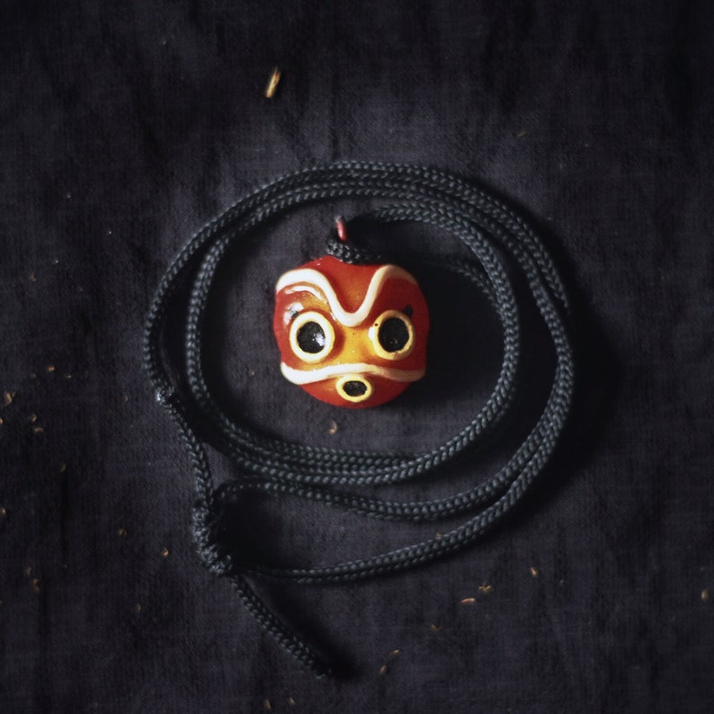 May include: A red and white mask with black eyes on a black cord necklace. The mask has a white outline and a red mouth.