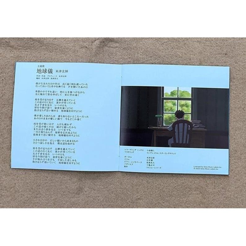 May include: A blue book with Japanese text on the left page and a black and white illustration of a person sitting at a desk looking out a window on the right page. The text on the left page includes the title "地球儀" which translates to "Globe".