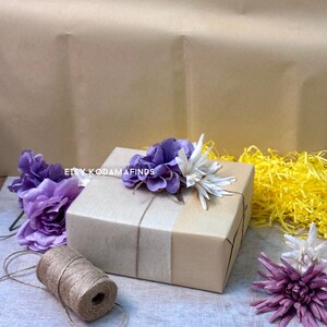 May include: A brown gift box wrapped with white tissue paper and tied with twine. The box is decorated with purple and white artificial flowers. The text "Etsy Kodamafinds" is visible on the box.