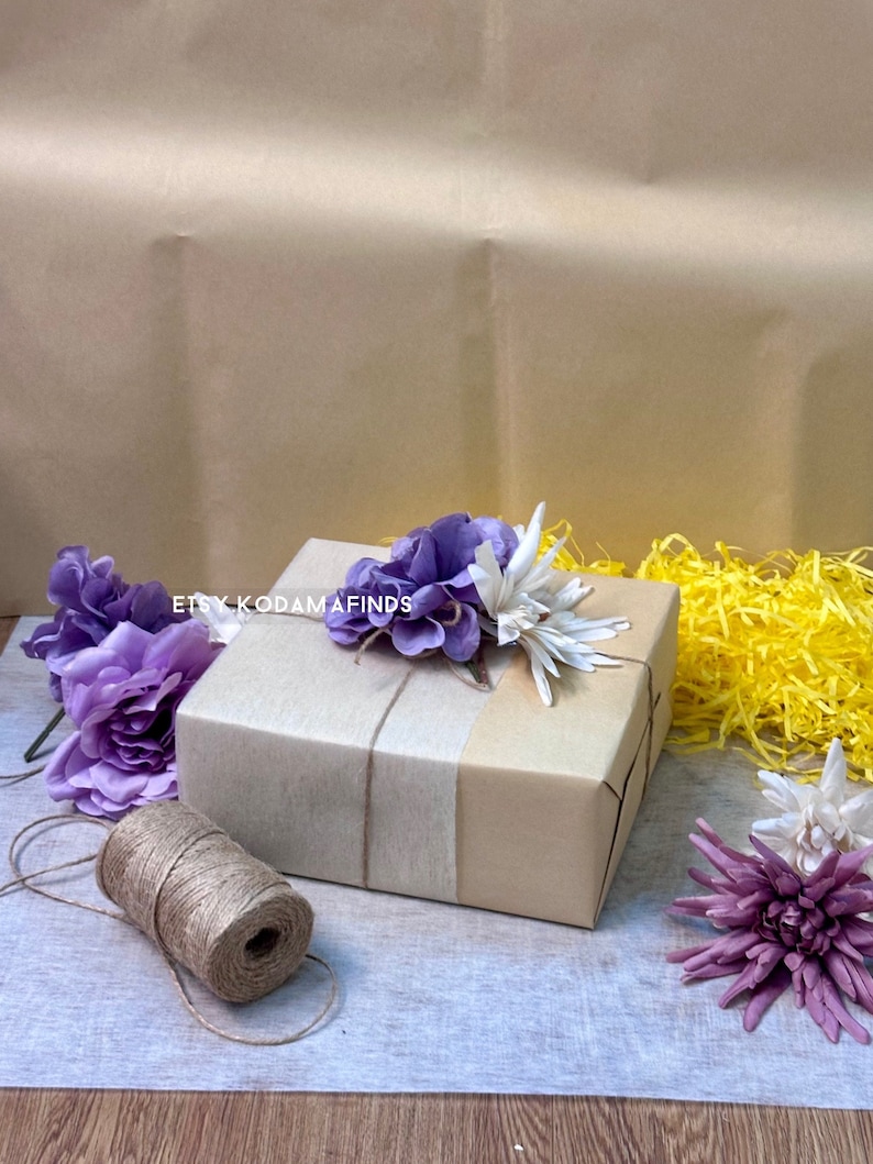 May include: A brown gift box wrapped with white tissue paper and tied with twine. The box is decorated with purple and white artificial flowers. The text "Etsy Kodamafinds" is visible on the box.