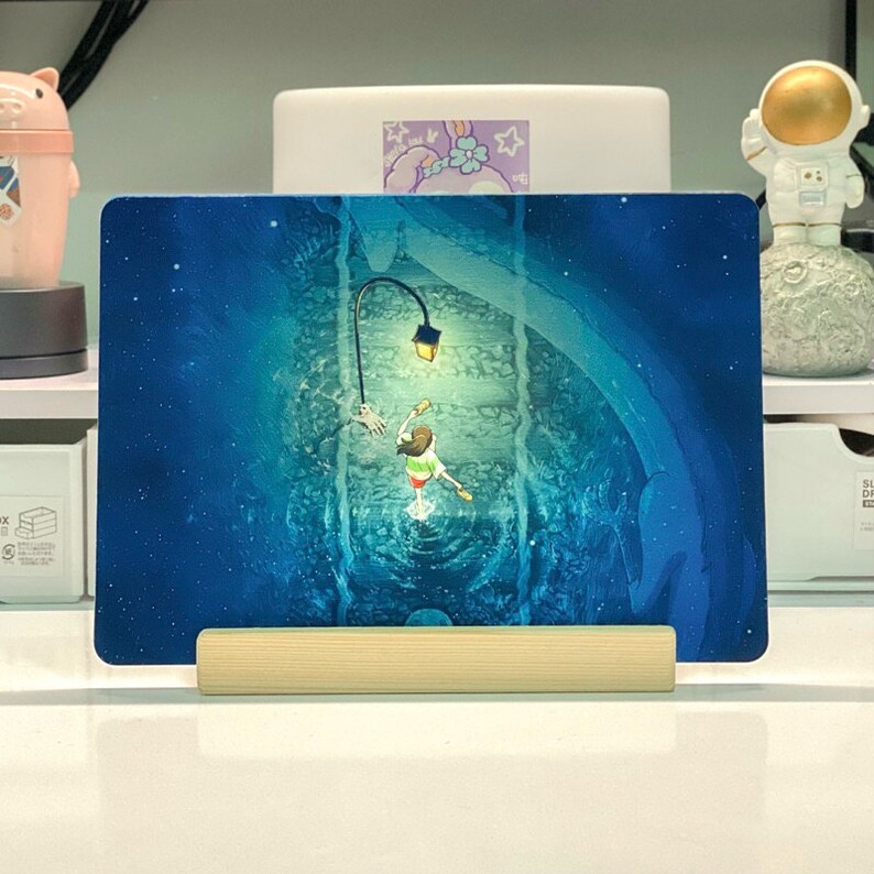 May include: A blue and white postcard with a scene of a young girl walking on a path with a streetlight. The girl is wearing a green shirt and a blue skirt. The path is surrounded by water and the background is a dark blue sky with stars.